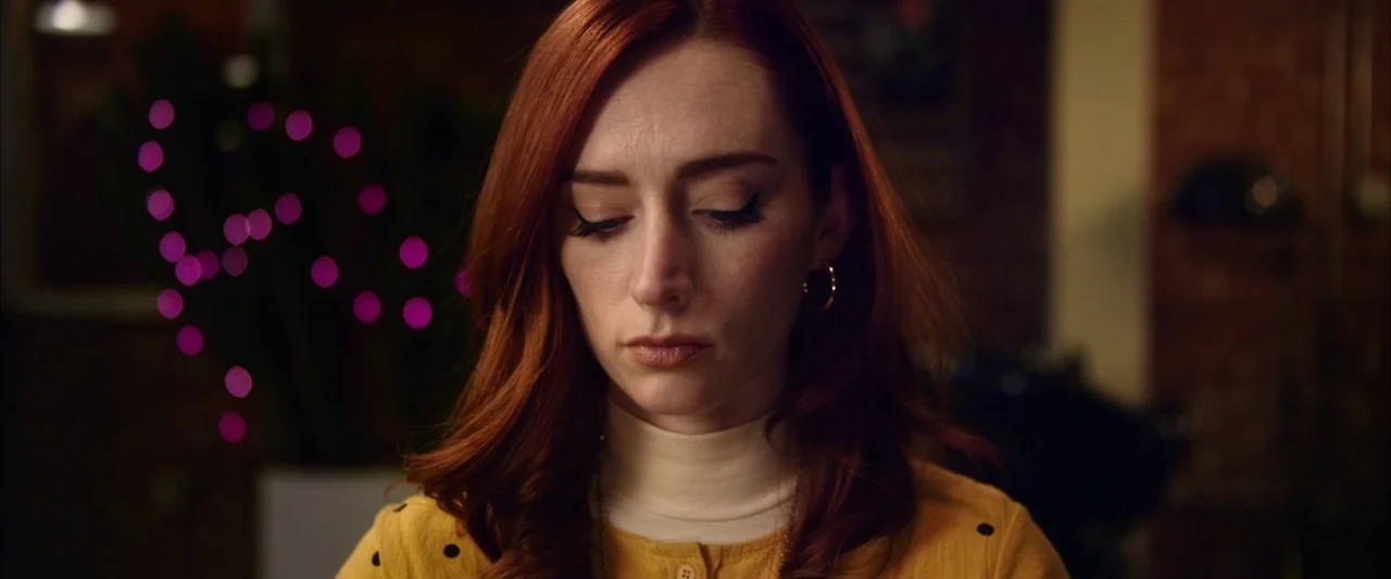 The Stylist (2020) movie still: closeup — A woman with red hair sits indoors against a blurry background of purple bokeh lights. She gazes downward…