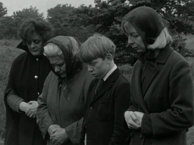 Children (1976) movie still: medium — Four people stand in a grassy cemetery during a muted, overcast day. The group bows their heads collectively…