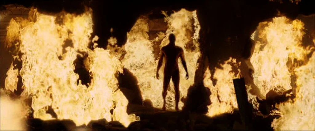 V for Vendetta (2006) movie still: fullbody — A dark, charred landscape is engulfed by tall, intense flames on all sides. A singular, dark, hairless…