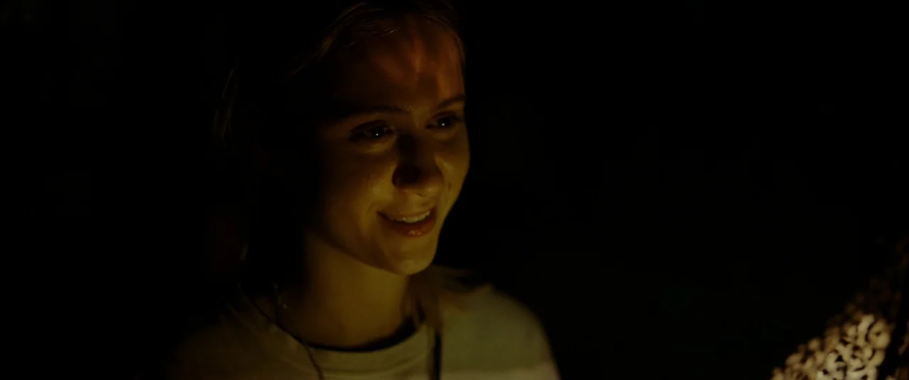 Bodies Bodies Bodies (2022) movie still: closeup — A woman's face is illuminated in a dark room by an off-screen light. The light casts soft warm…
