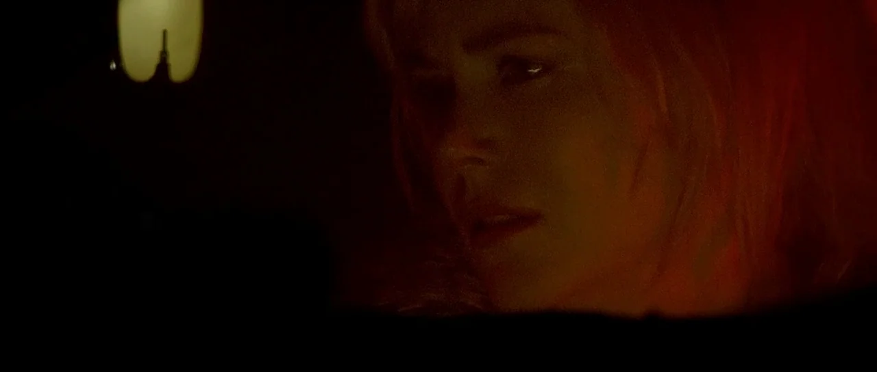 Dogville (2003) movie still: closeup — A dark indoor setting is lit only by a faint, warm light source. A woman with light hair is partially visible…