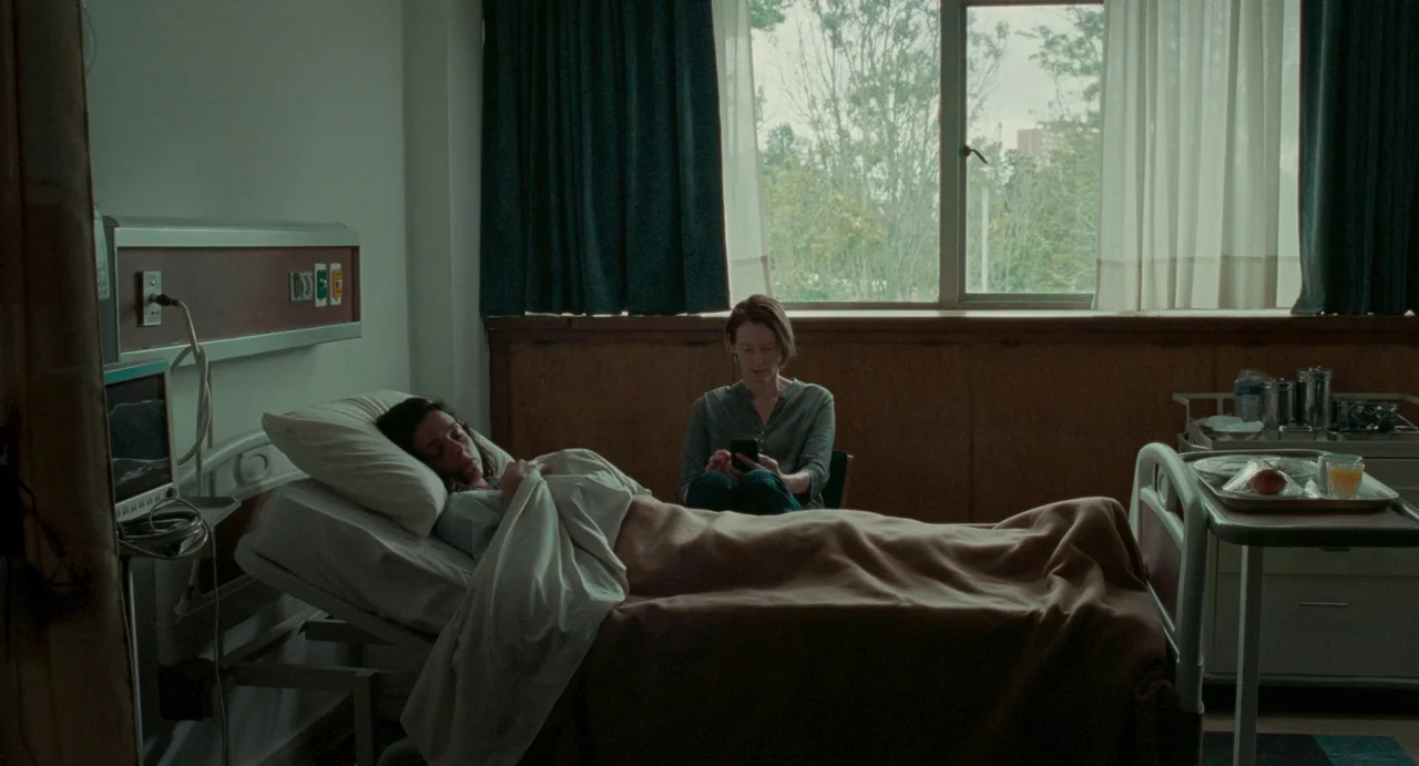 Memoria (2021) movie still: wide — A hospital room features a bed positioned in front of a window with curtains. A woman rests in the bed while…