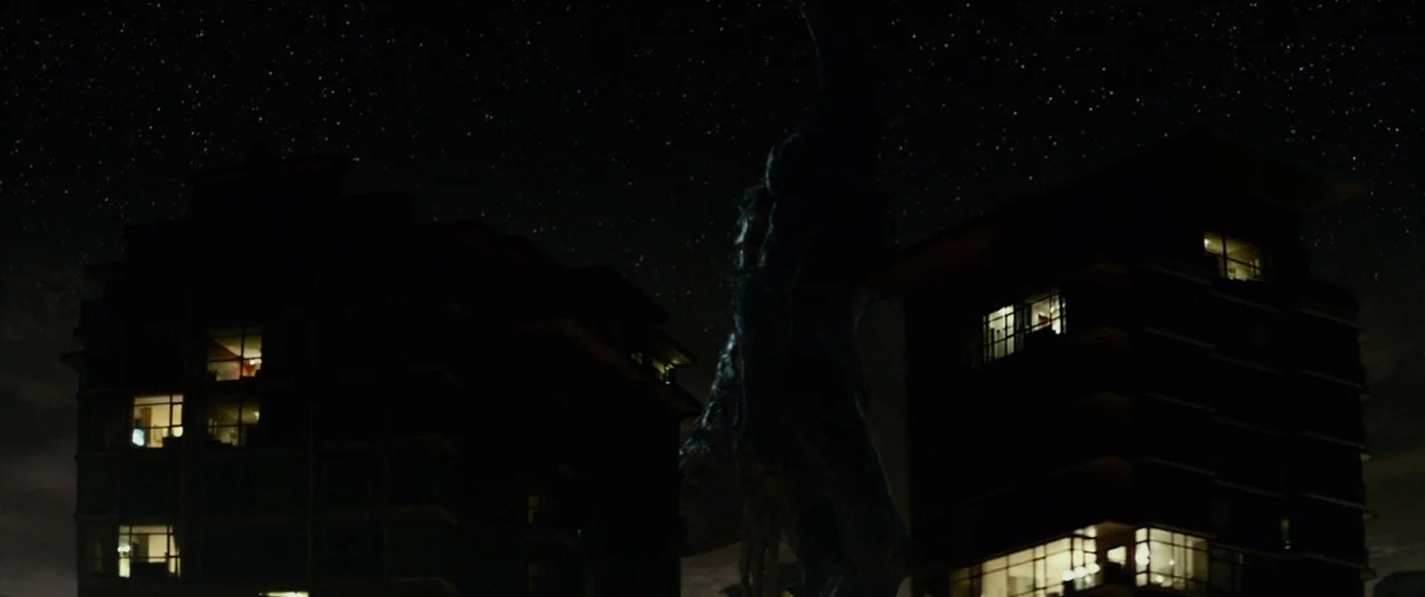 Colossal (2016) movie still: wide — A monolithic creature stands between two darkened residential buildings under a starlit night sky. The creature…