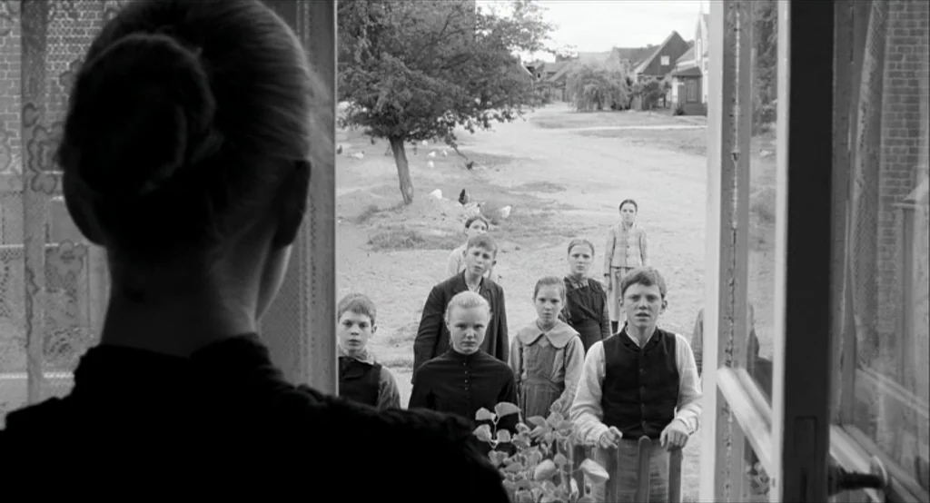 The White Ribbon (2009) movie still: medium — A rural village setting with a group of children gathered outside a wooden doorway. The children wear…