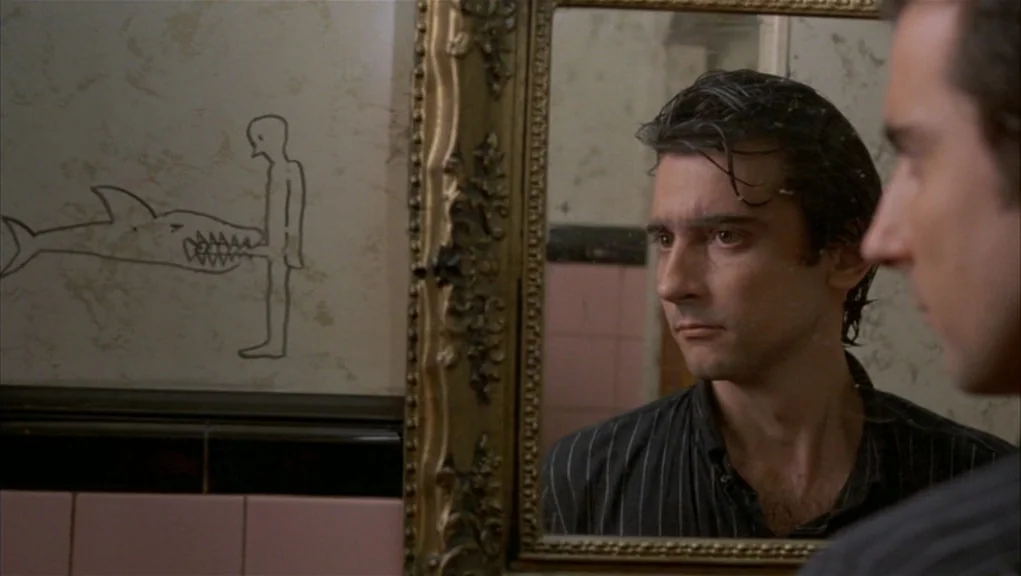 After Hours (1985) movie still: medium — An interior bathroom features peeling wallpaper and a framed rectangular mirror above pink tiles. A man…