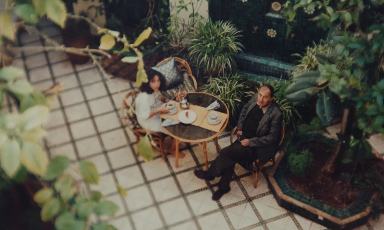 Little Girl Blue (2023) movie still: wide — An tiled patio features a small glass-topped table and leafy potted plants. A man and a woman sit at the…