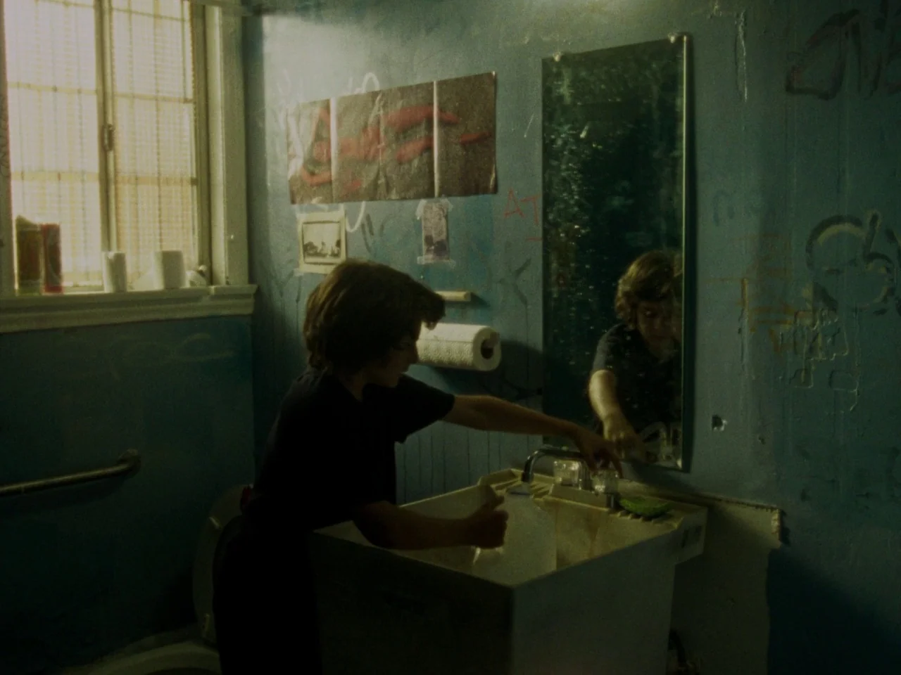 Mid90s (2018) movie still: medium — A dimly lit room with blue walls covered in faded posters and graffiti. A young person washes a glass under the…