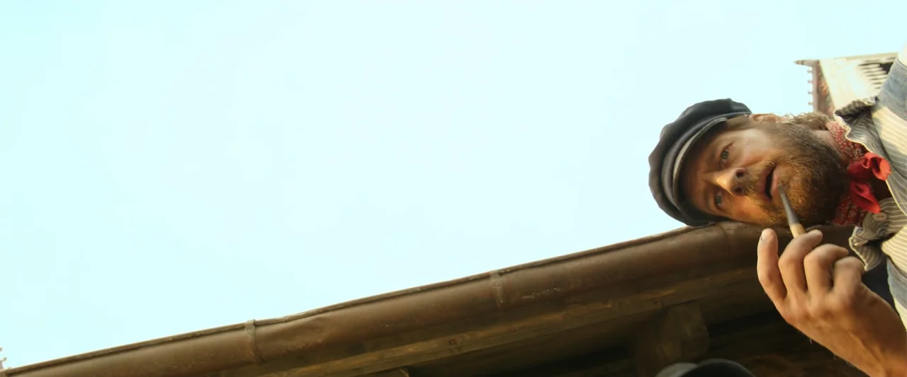 Jim Button and Luke the Engine Driver (2018) movie still: medium — A man stands near the edge of a wooden roof structure against a clear sky. He…