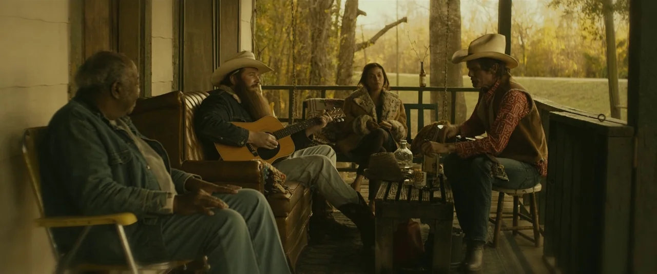 Blaze (2018) movie still: wide — A wooden porch overlooks a rural landscape during the soft light of golden hour. Four adults are gathered together,…