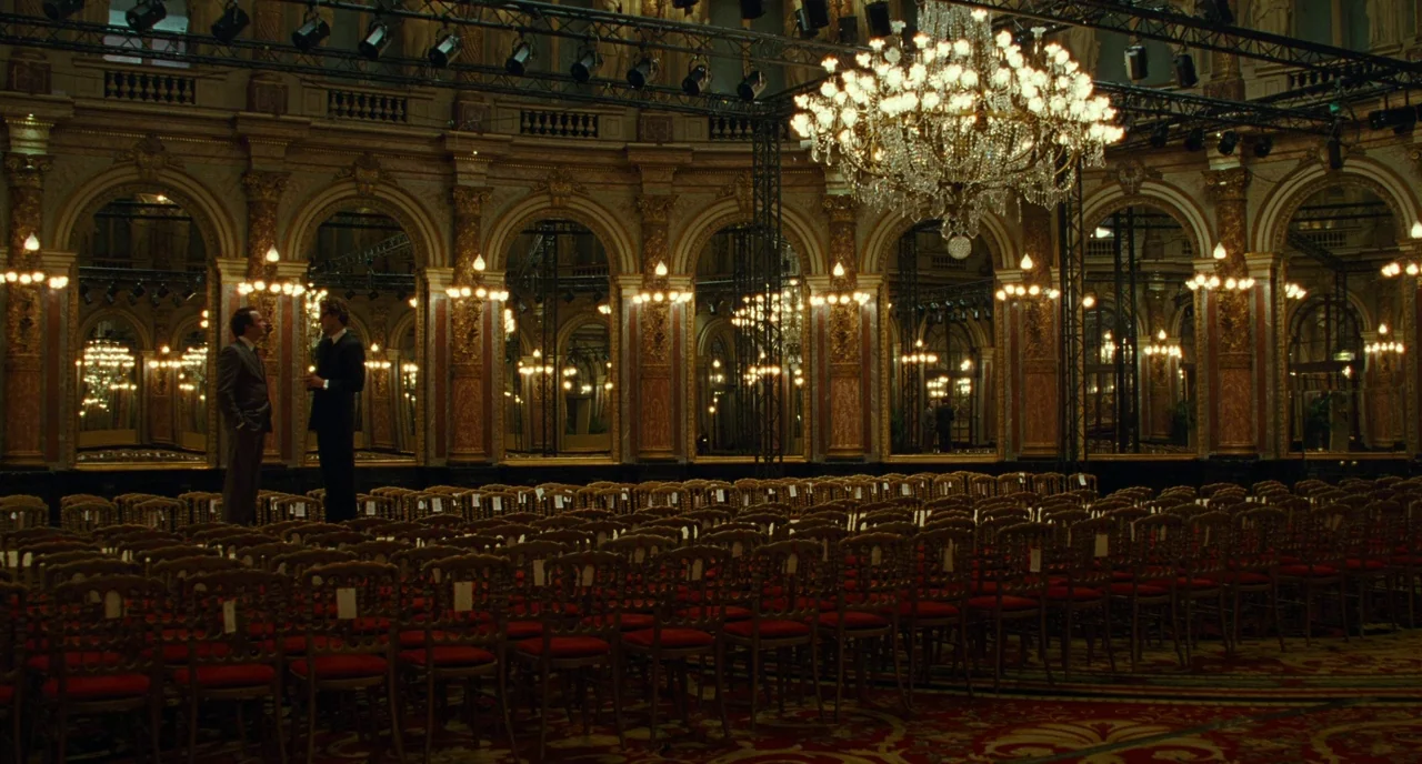Saint Laurent (2014) movie still: wide — An opulent ballroom features high arched mirrors, golden decorative trim, and a large central chandelier.…