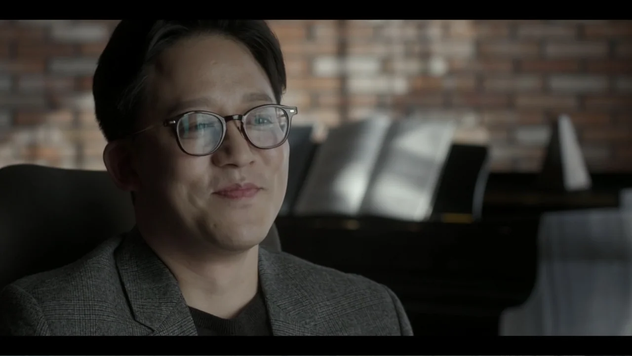 Lee Soo Man: King of K-Pop (2025) movie still: closeup — A man sits in a dimly lit space with a blurred brick wall in the background. The man wears…