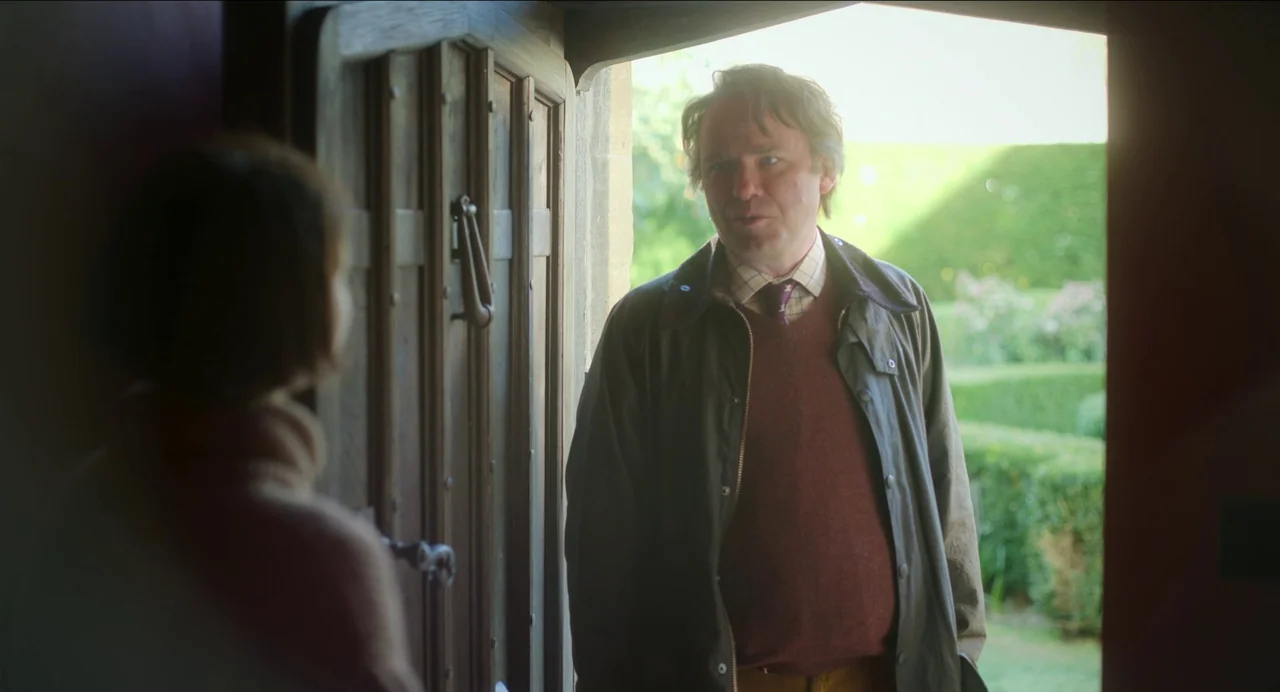 Men (2022) movie still: medium — A man stands in the doorway of a stone house looking outside at someone. Sunlight illuminates the man from behind…