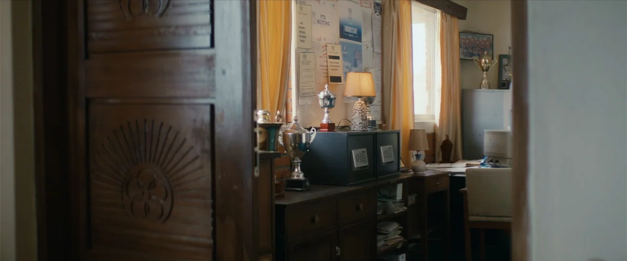 Ms. Kanyin (2025) movie still: medium — A cluttered school office features wooden furniture, metal safes, and various trophies on display. Sunlight…