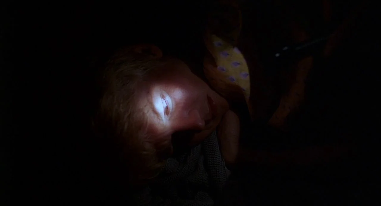 Psycho III (1986) movie still: closeup — A dark, nondescript interior space at night. A man lies down with his face illuminated by a direct beam of…
