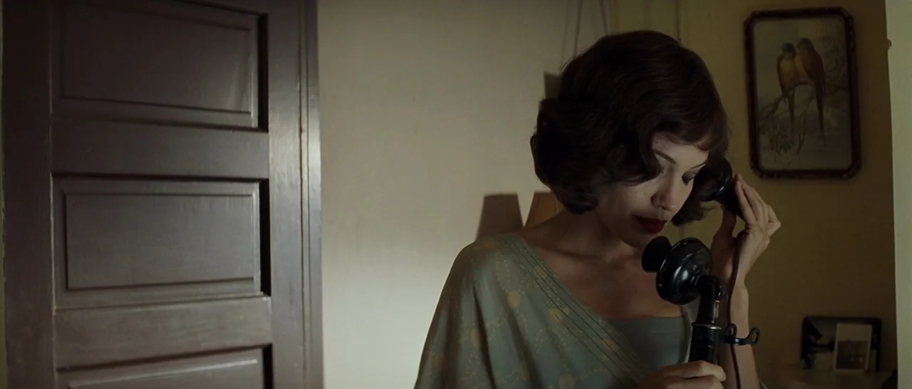 Changeling (2008) movie still: medium — A dimly lit indoor space with a plain wall and a closed wooden door. A woman with dark wavy hair holds a…
