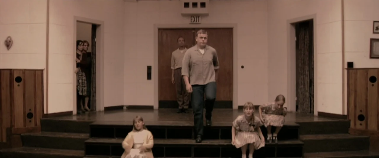 Dancer in the Dark (2000) movie still: fullbody — A wooden stage in a room with white paneled walls and multiple doors. A man walks down the steps…