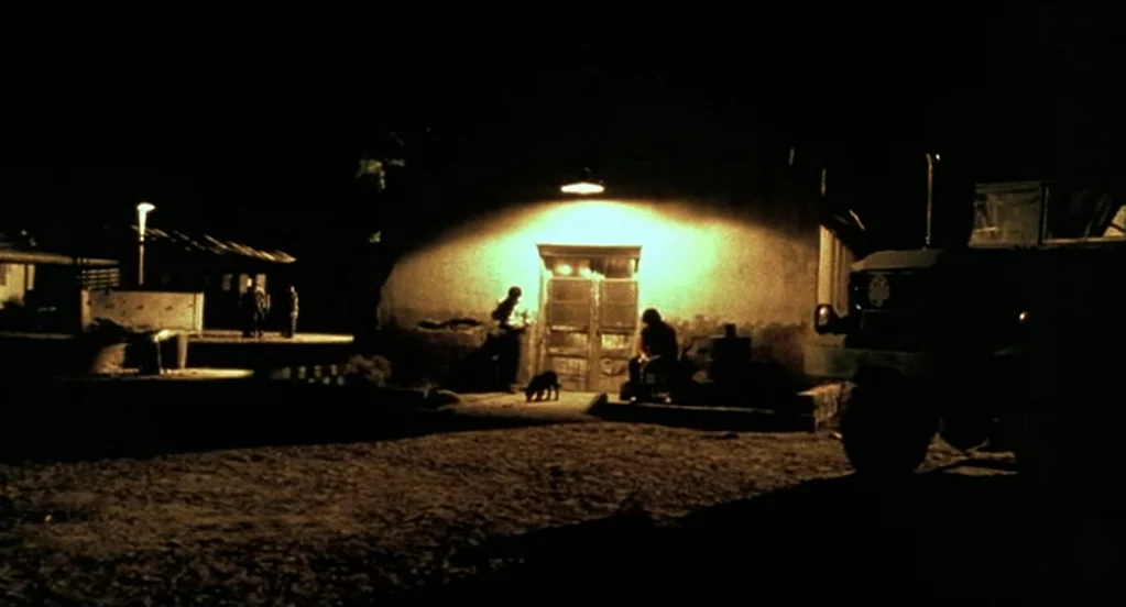 Traffic (2000) movie still: wide — A dark outdoor area features a single lit doorway on a plain building wall. Two men sit near the entrance while a…
