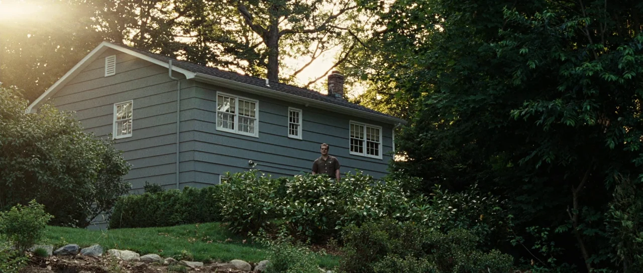 Revolutionary Road (2008) movie still: wide — A suburban house with grey siding stands amidst dense green trees and bushes. A lone man stands in the…