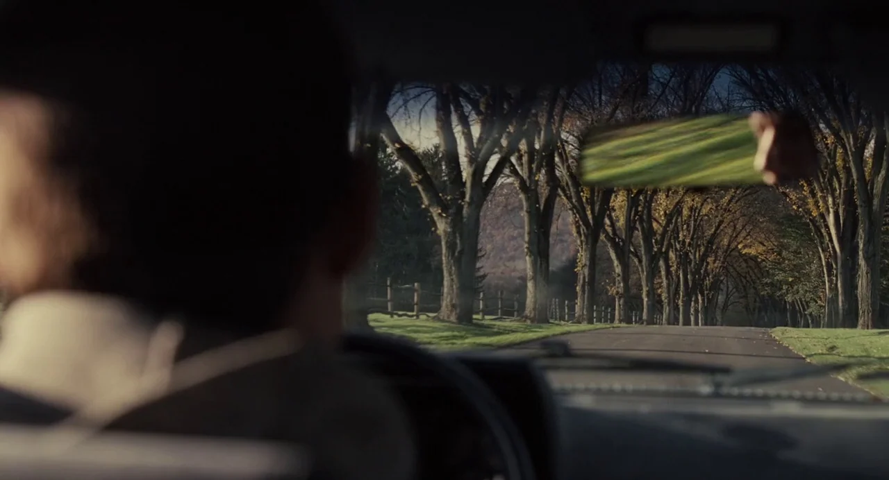 Foxcatcher (2014) movie still: medium — A view from inside a car looking out at a road lined with trees. A driver is visible in the foreground as…