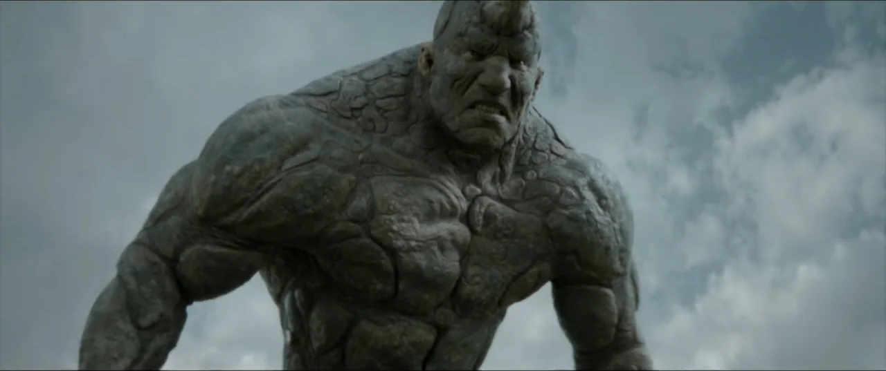 Kraven the Hunter (2024) movie still: medium — A massive, muscular creature with stone-like skin stands against an overcast sky. The figure stares…