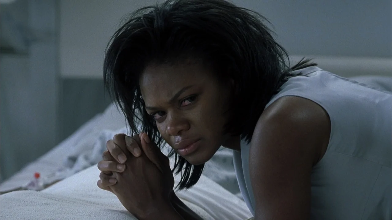 John Q (2002) movie still: closeup — A dark bedroom interior features a woman kneeling on a white mattress. She clasps her hands together tightly…