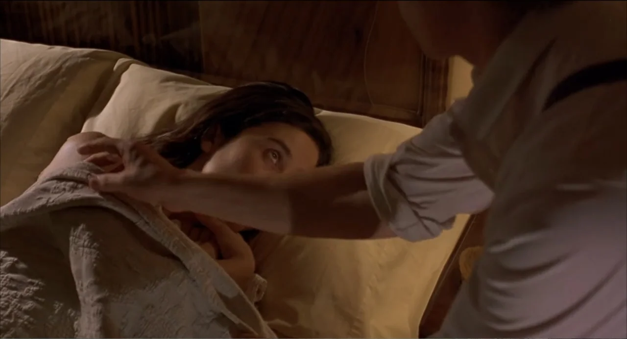 Like Water for Chocolate (1992) movie still: medium — A dimly lit bedroom features a wooden headboard and a person lying in bed. A man in a white…