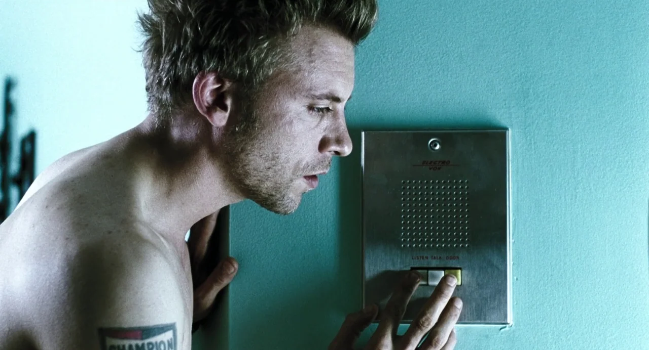 Last Night (1998) movie still: medium — A man stands against a smooth turquoise wall with a mounted metallic intercom panel. The man is pressing a…