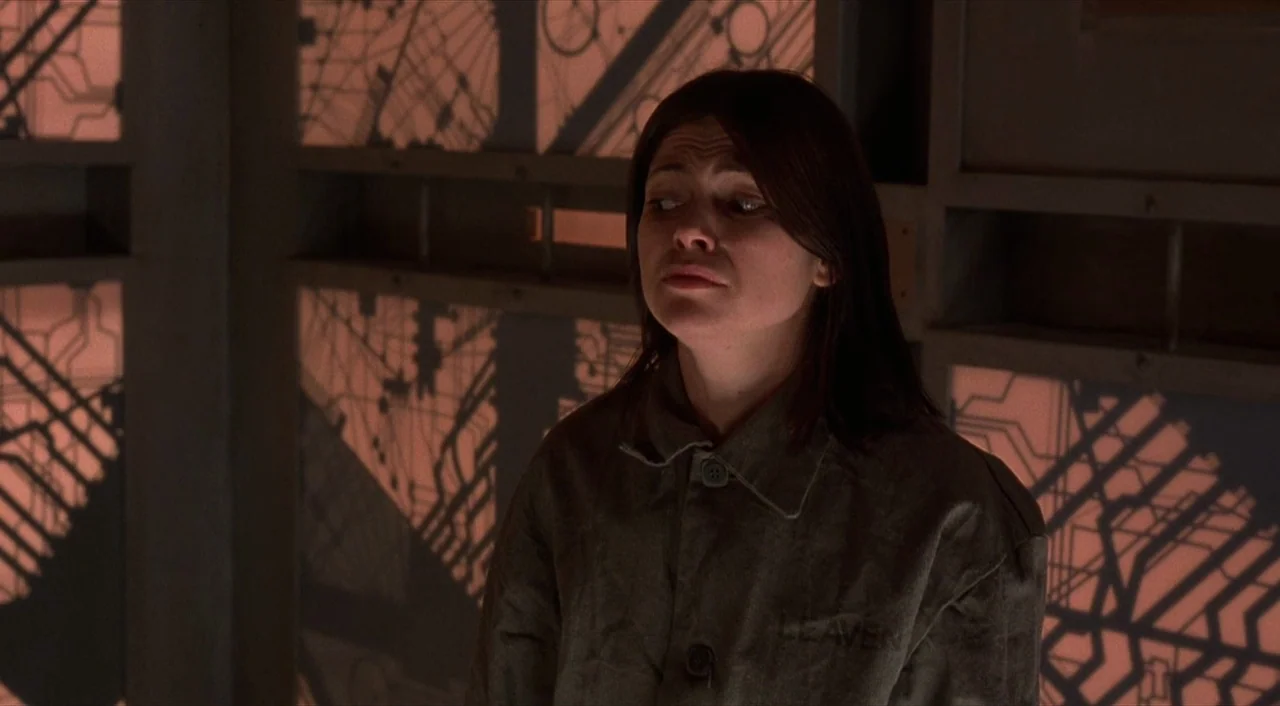 Cube (1997) movie still: medium — A woman stands in a claustrophobic enclosure with glowing, geometric circuit-patterned wall panels. She wears a…