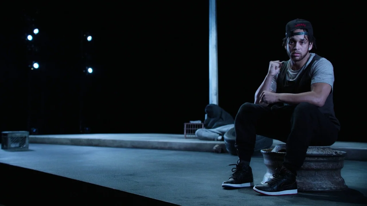 Pass Over (2018) movie still: medium — A minimalist studio set featuring a dark stage floor under artificial lighting. A man sits on a tire rim…