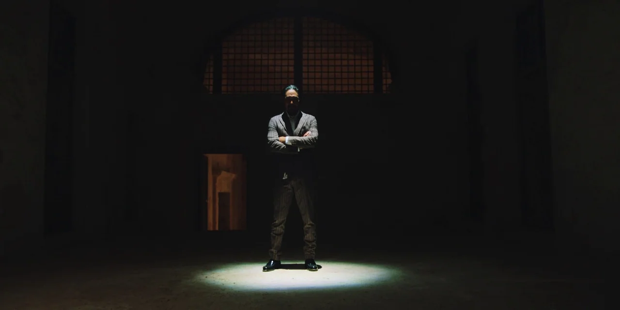 Sniper: G.R.I.T. (2023) movie still: fullbody — A dark warehouse interior with a large arched barred window in the background wall. A man in a…