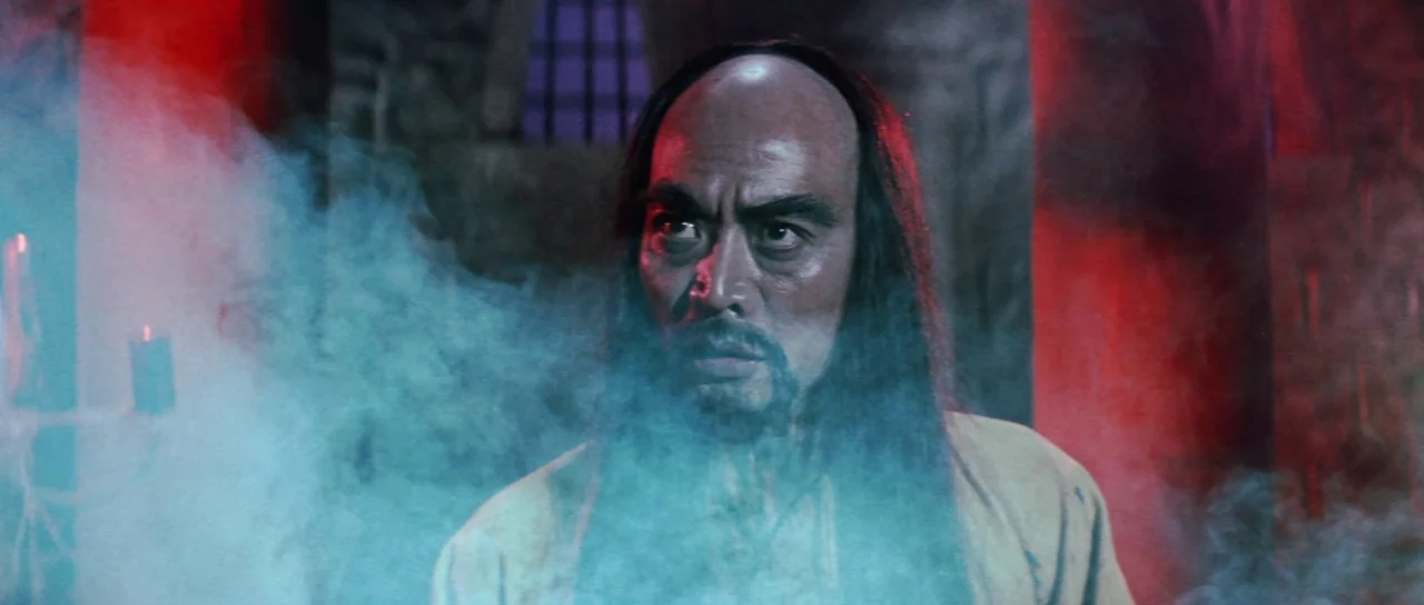 The Legend of the 7 Golden Vampires (1974) movie still: closeup — An older man stands inside a dimly lit stone chamber filled with colored smoke. He…