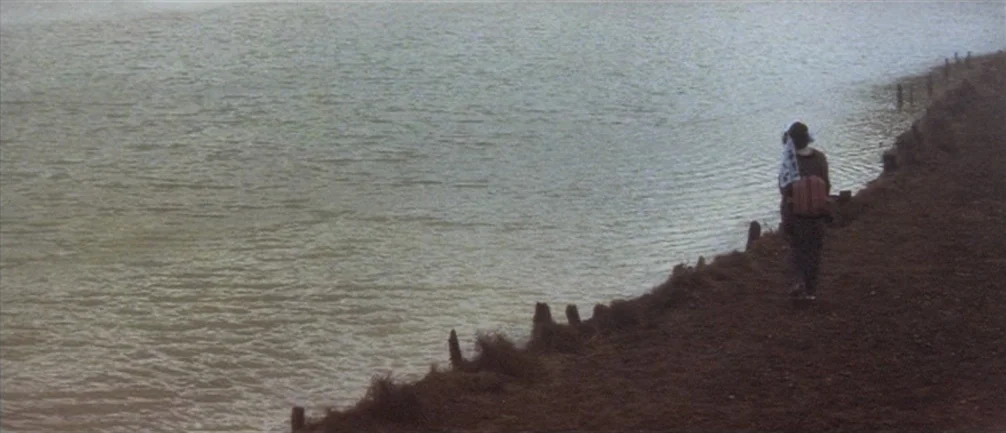 An Actor’s Revenge (1963) movie still: wide — A rural lakeshore features dark, textured earth and a row of low wooden posts. A single person wearing…