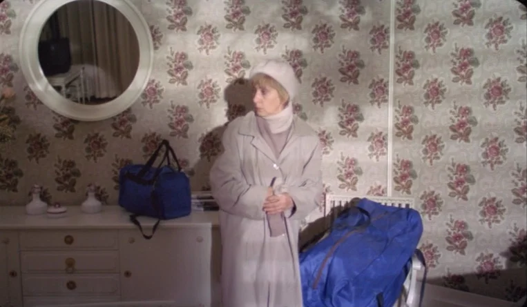 The Turn of the Century (2001) movie still: fullbody — A room features floral patterned wallpaper and a white wooden dresser with two bags. A woman…