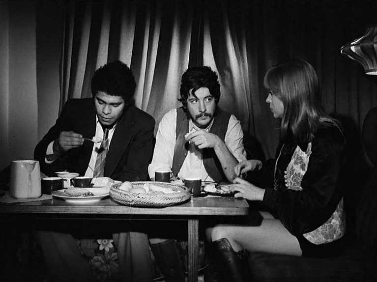 Gods Of The Plague (1970) movie still: medium — Three people sit at a small table inside a dimly lit booth. They are eating a meal together in a…