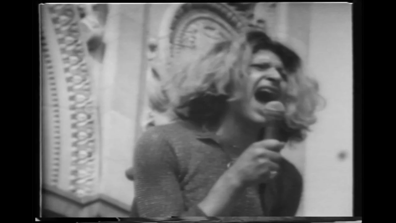 Orlando: My Political Biography (2023) movie still: medium — A person stands in front of an ornate stone building facade. The individual sings…