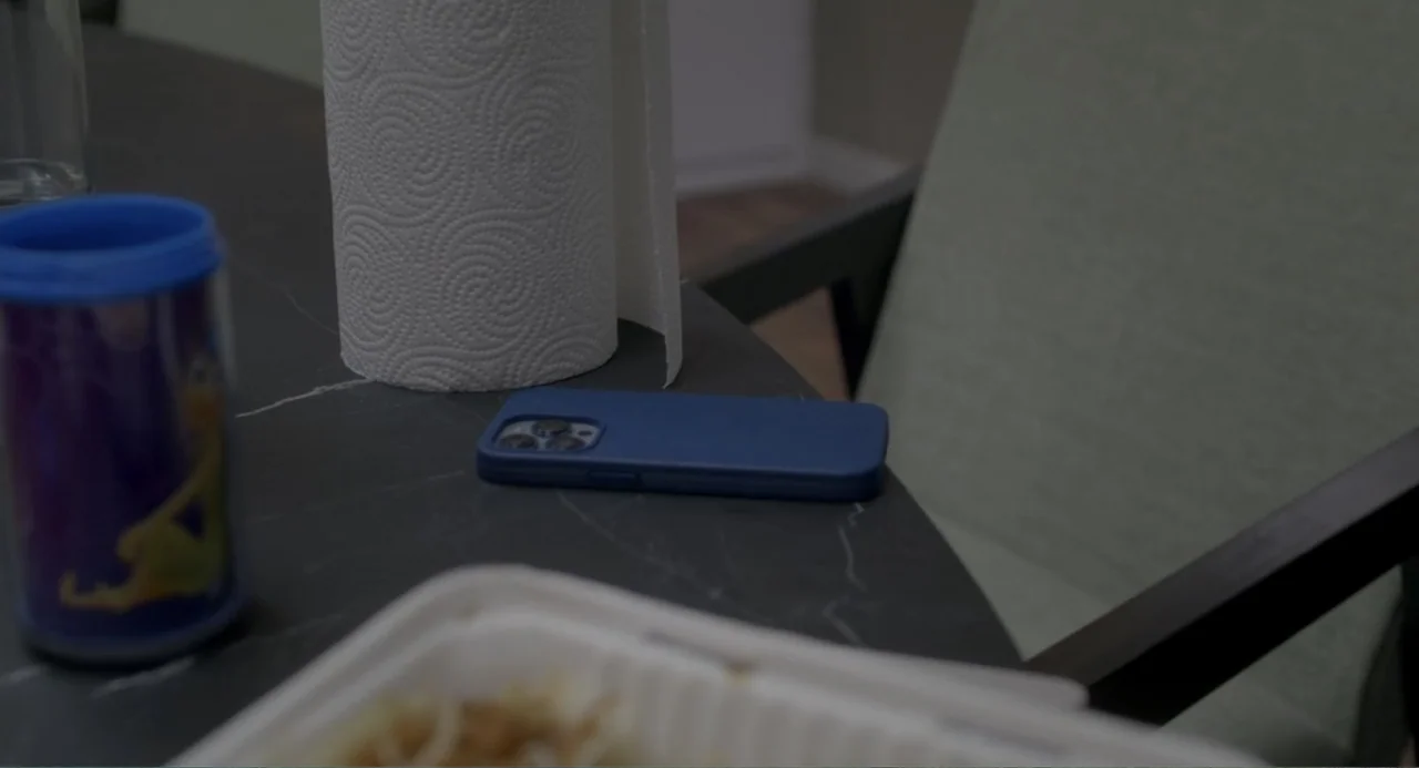 Straight (2024) movie still: medium — A close overhead view of a dark marble surface with dining items. A blue smartphone, a roll of paper towels, a…
