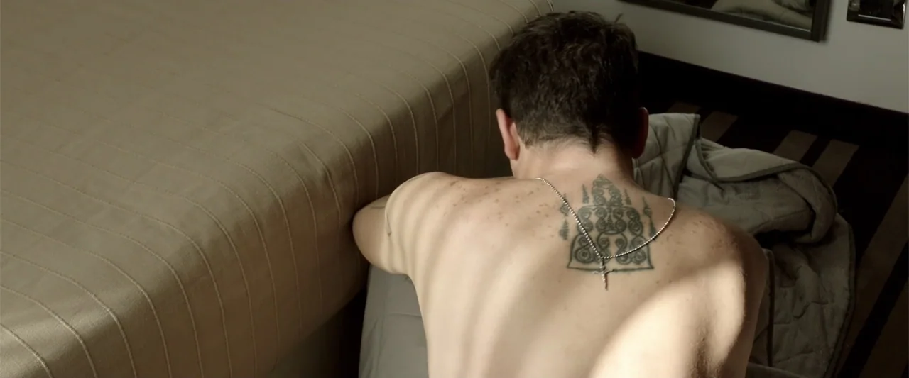 About Us (2017) movie still: medium — A spare modern hotel room features a simple beige bedspread and a striped rug. A man with a tattoo on his back…