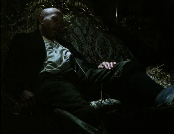 The Tempest (1979) movie still: medium — A dark, cluttered barn interior featuring an ornate, antique high-backed chair filled with straw. A bald…