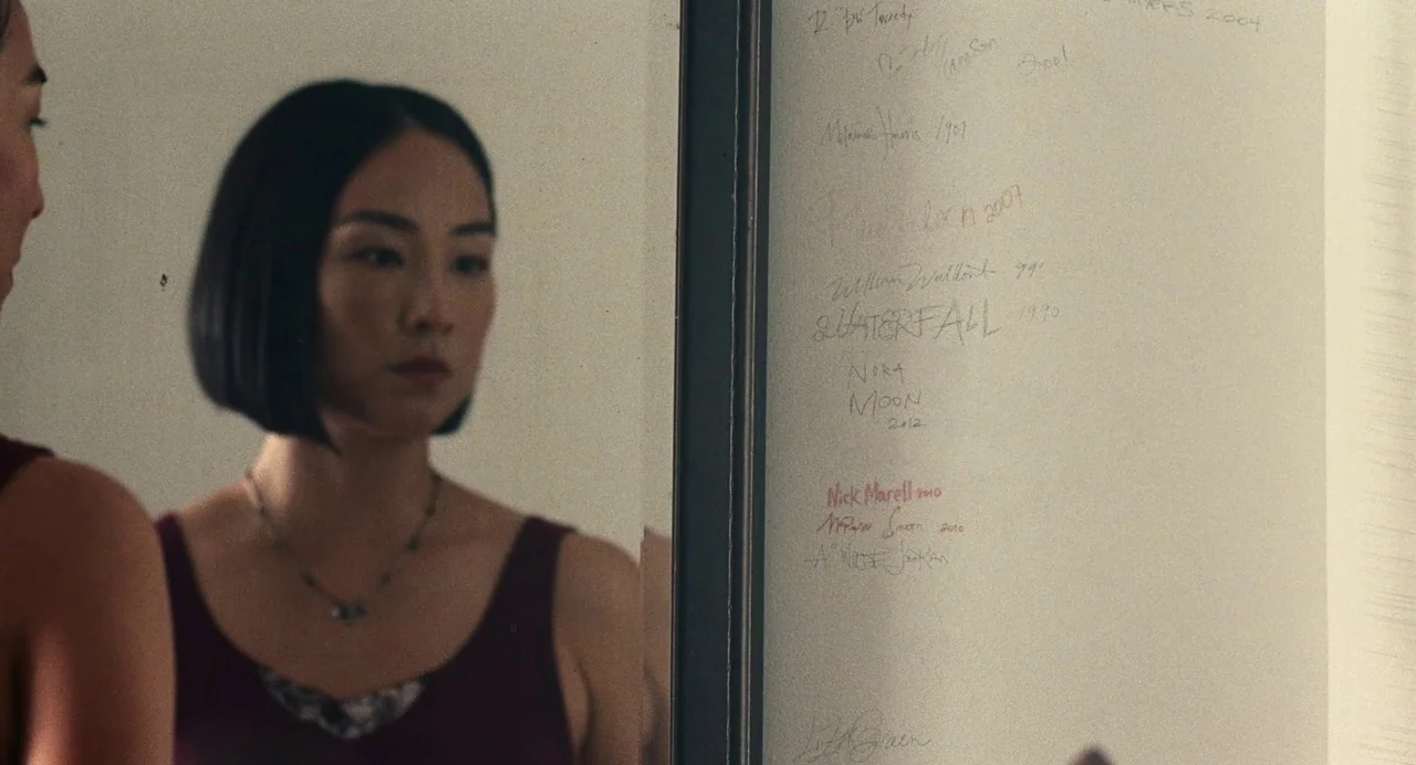 Past Lives (2023) movie still: medium — A woman stands before a wall covered in various handwritten names and years. She stares at her own…