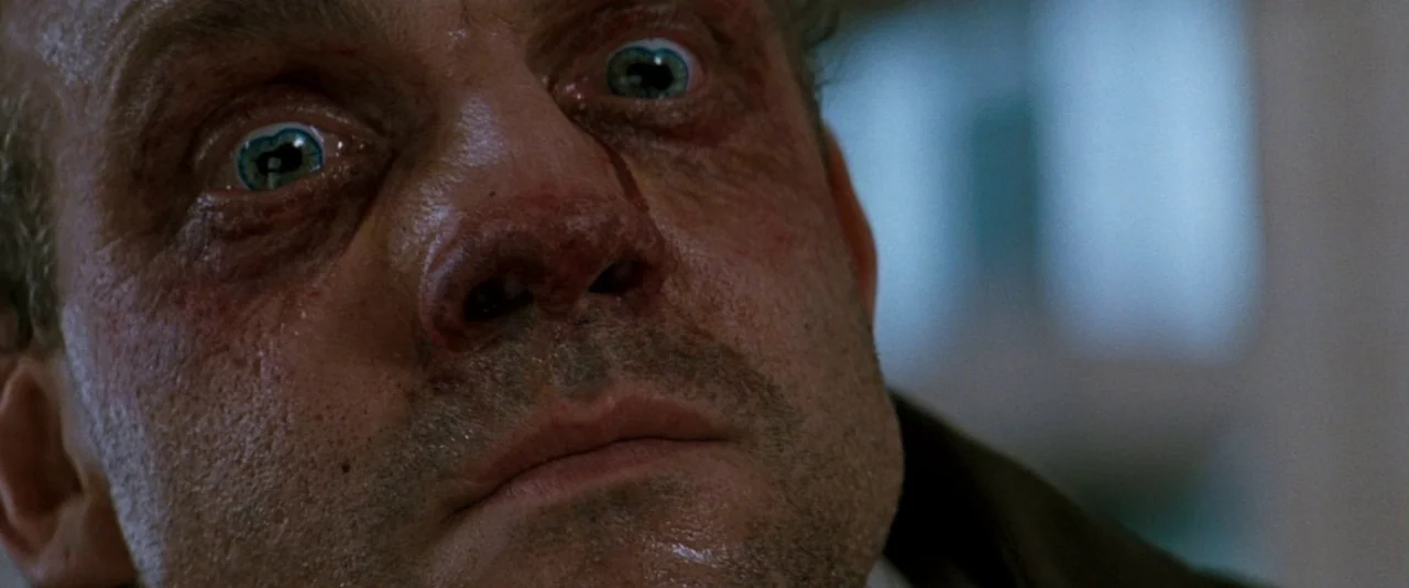 In The Mouth of Madness (1994) movie still: closeup — A close view of a man's face, showing skin pores and light sweat. The subject stares directly…