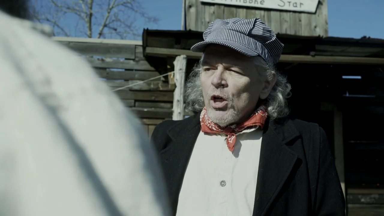 The Dalton Gang (2020) movie still: medium — An older man in western attire stands outdoors in front of a rustic wooden building. The man speaks…