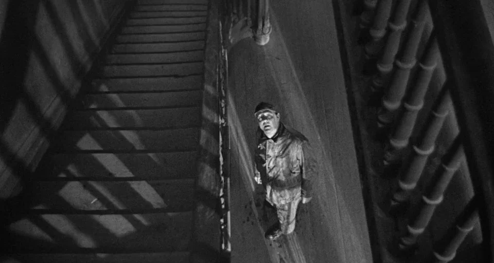 Baby Doll (1956) movie still: wide — A dimly lit staircase features dramatic shadows cast by a wooden banister. A man stands on the ground floor…