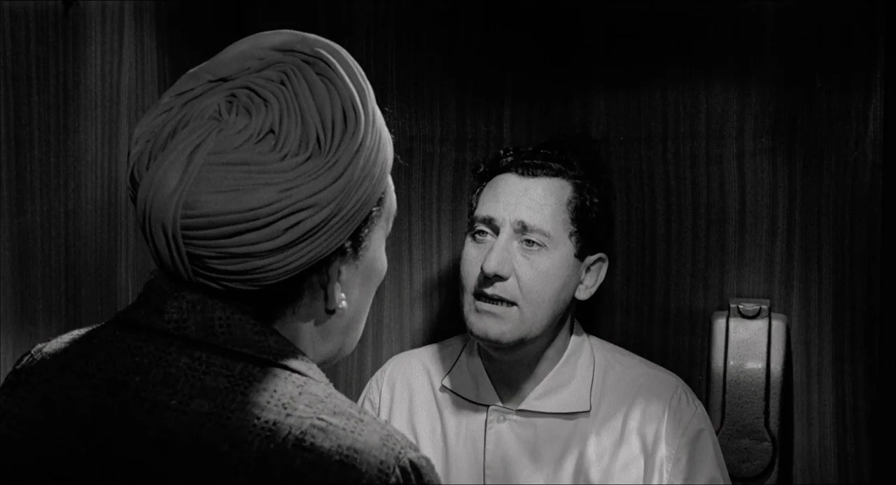 Il Boom (1963) movie still: medium — Two individuals occupy a tight wooden space against a dark, paneled wall. A man faces a woman while speaking,…