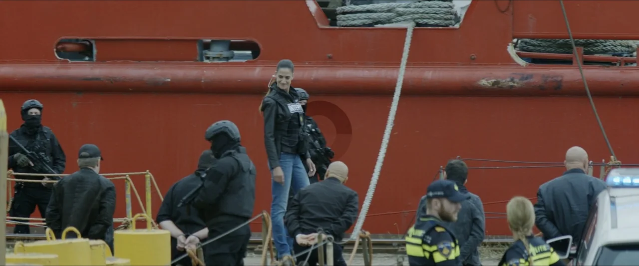 Black Lotus (2023) movie still: fullbody — A group of police officers and handcuffed individuals stands before a large red ship hull. A woman in…