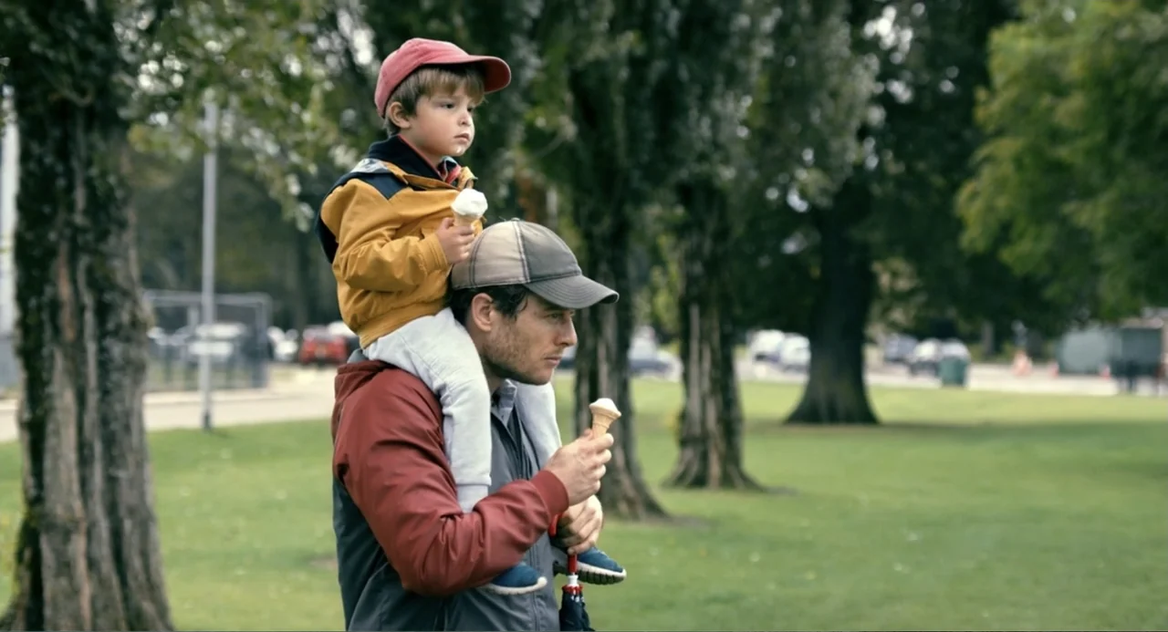Living Out Loud (1998) movie still: medium — A green public park with several tall trees in the background during daylight hours. A man with a child…
