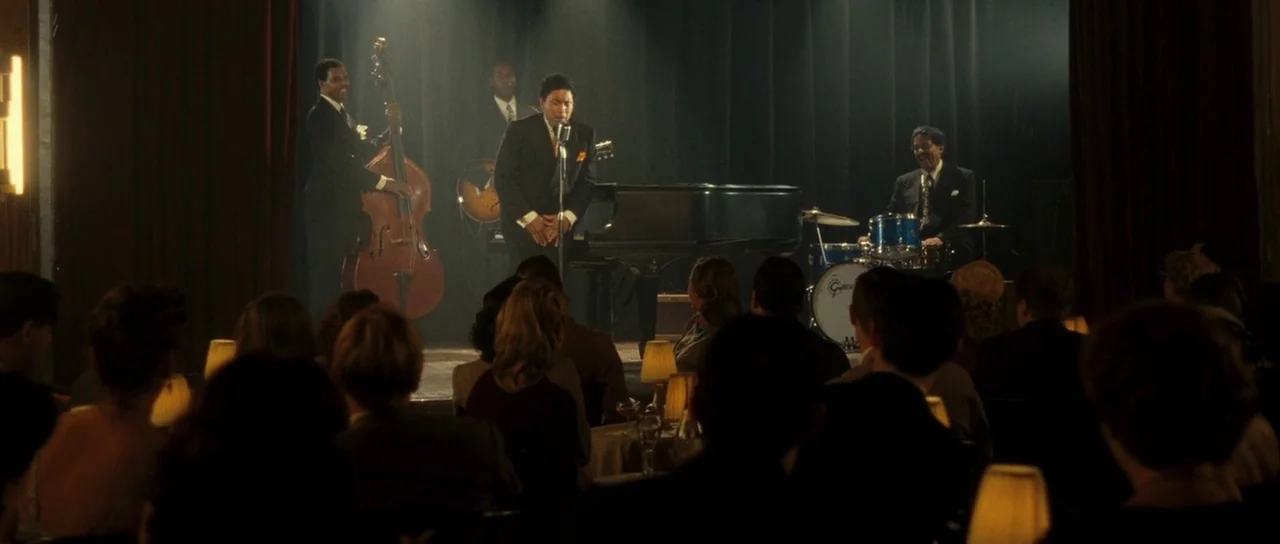 On The Road (2012) movie still: wide — A dimly lit club with a jazz quartet performing on a curtained stage. Patrons sit at round tables in the…
