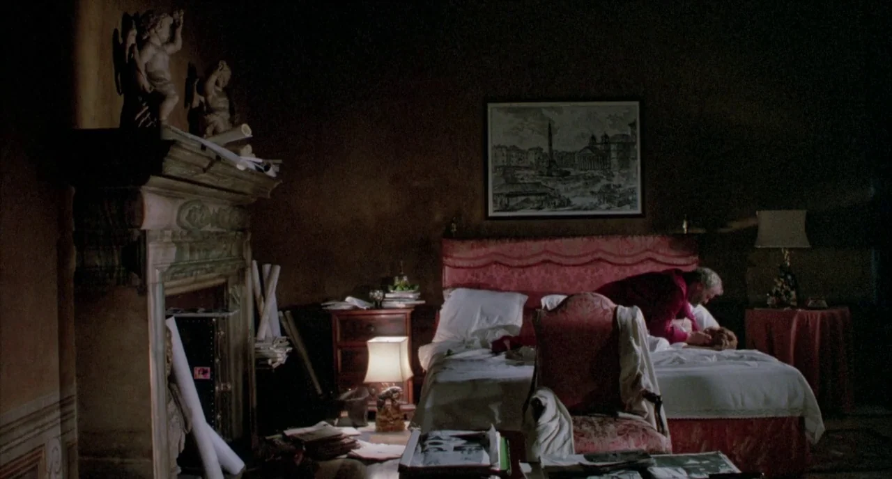 The Belly of an Architect (1987) movie still: wide — A dimly lit room contains a large bed with a crimson headboard and a fireplace. A man in a dark…