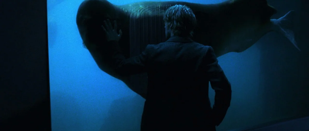 Stay (2005) movie still: medium — A man in a dark suit stands before a large, illuminated water tank. He places his right hand against the glass…