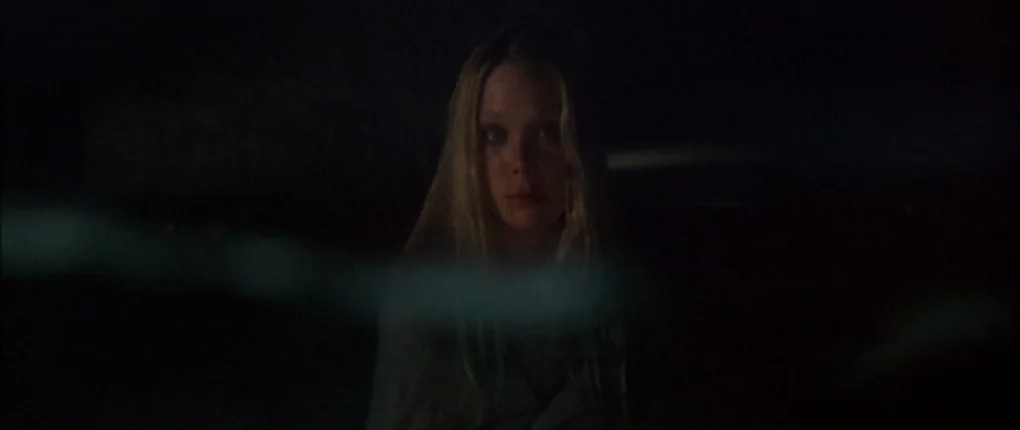 3 Women (1977) movie still: closeup — A dark, nondescript outdoor area at night with a blurry foreground obstruction. A young woman with long blonde…