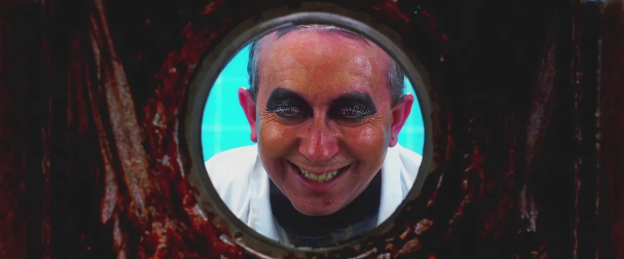 Excision (2012) movie still: closeup — A man peers through a circular metal window in a dark blood-covered door. He wears theatrical makeup and…